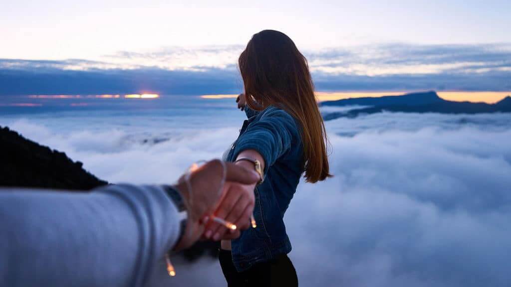 A couple stands together on a mountaintop, holding
hands and gazing out at the clouds below