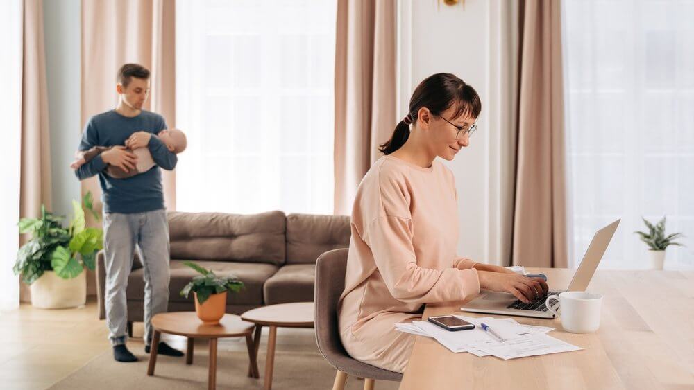 husband carrying a bay while wife is working on her laptop