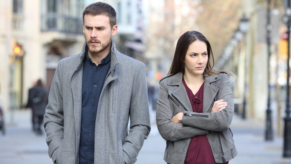 couple wearing grey coats standing on a street unhappily