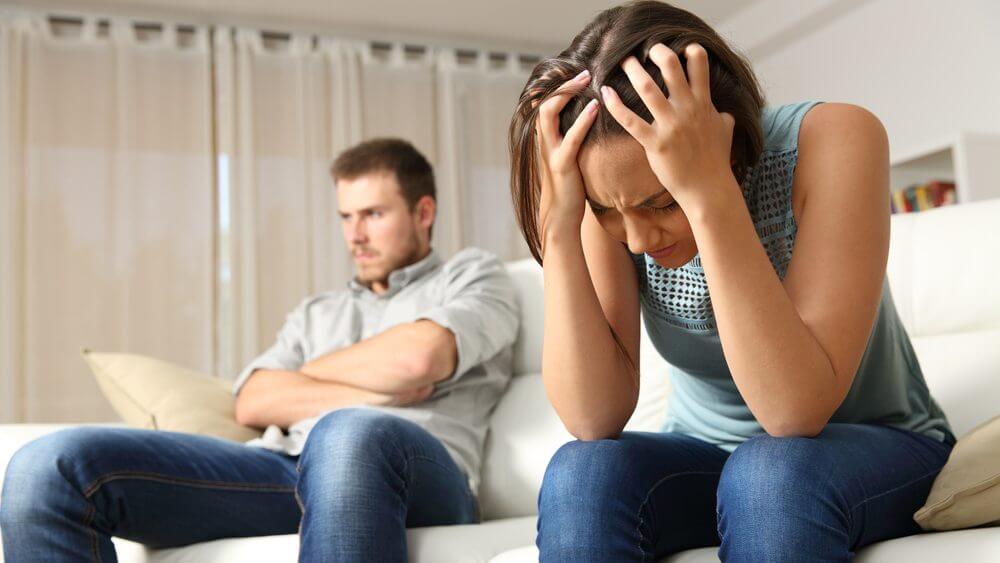 man sitting on white sofa with arms folded next to female partner with hands on head