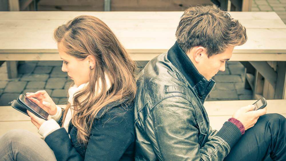 young couple in black jackets chatting on their phones with back against each other