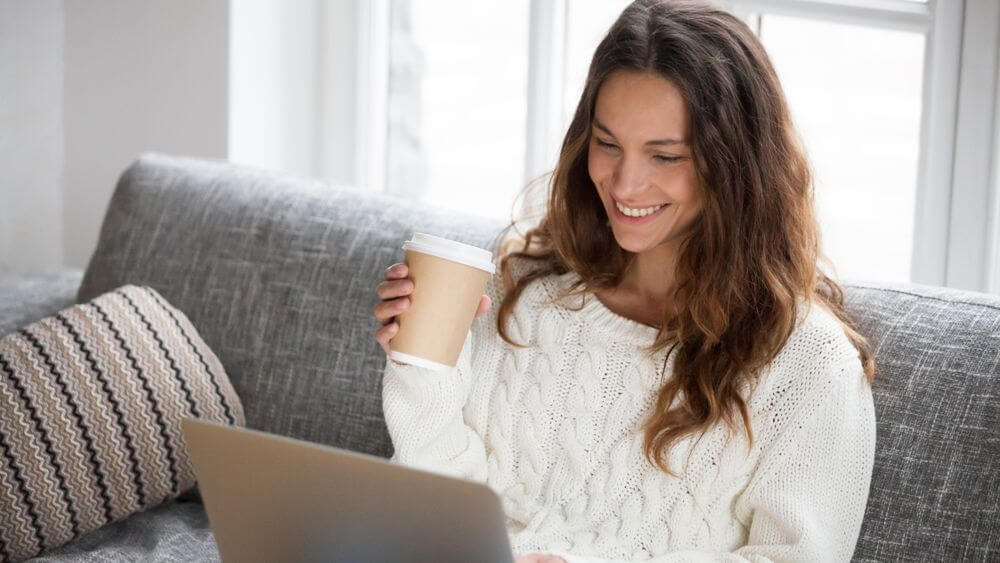 woman sitting on a grey sofa drinking coffee while video chatting on her PC