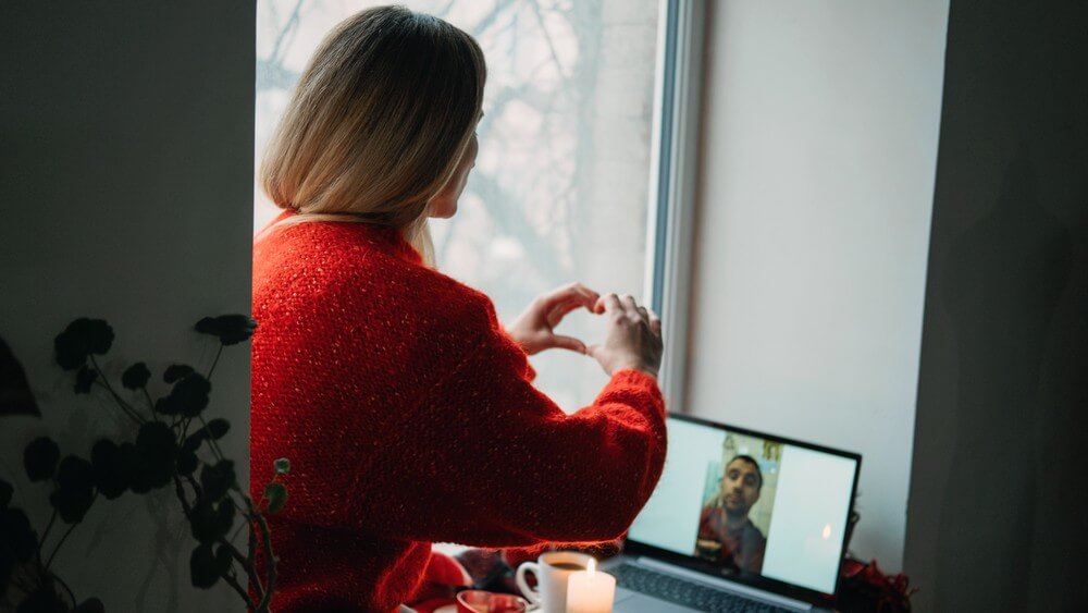 woman wearing red knitwear making a love symbol in a video call with her boyfriend