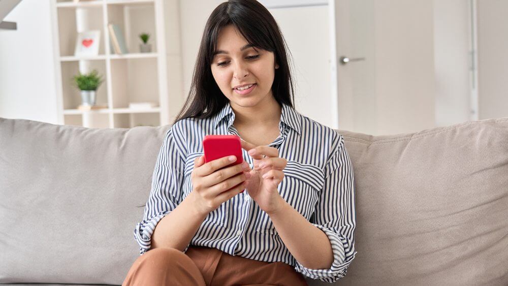 young beautiful Indian woman sitting on a sofa chatting with a red smartphone