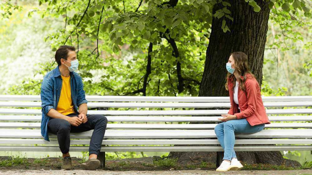 couple wearing masks, sitting apart on a white bench at a park