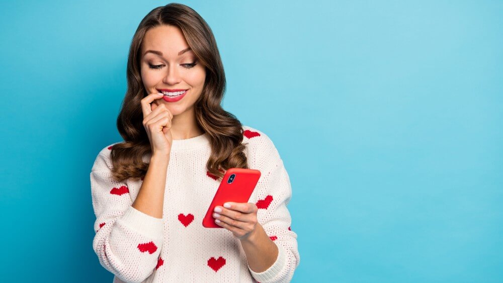 young woman wearing white knitwear with heart symbols on blue background, red smartphone