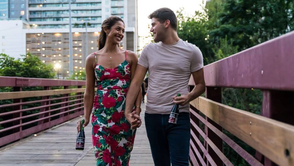 couple smiling, drinking, holding hands, and walking on a wooden bridge