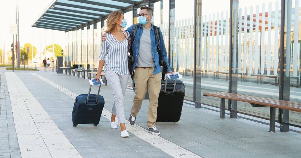 couple wearing masks and looking at each other rolling their luggage the airport terminal