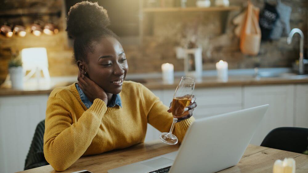 Young African American woman using laptop and toasting with Champagne while chatting on her PC